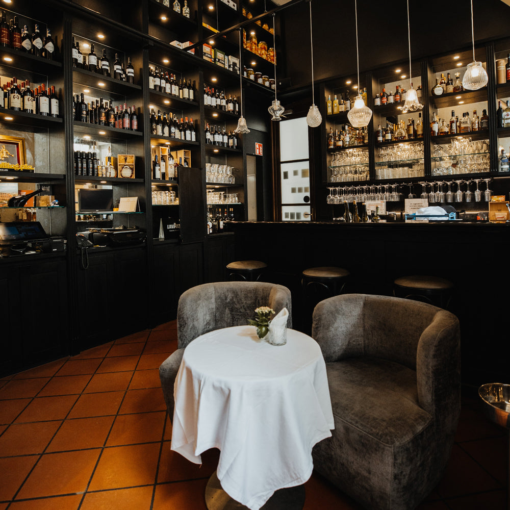 Aufnahme der Bar im Vordergrund ein Tisch mit zwei Stühlen im Hintergrund Regale mit Flaschen und Gläsern